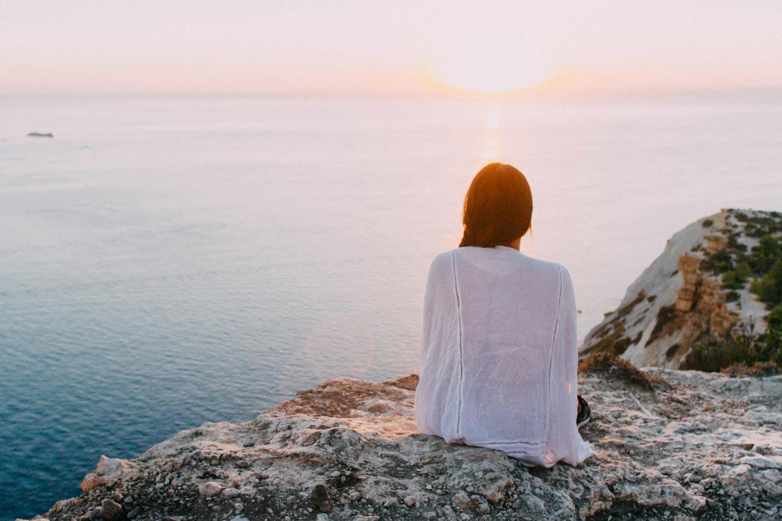 A serene view of a woman sitting on a rocky cliff, gazing at the ocean during sunset. Perfect for relaxation and travel themes. adult mental health services