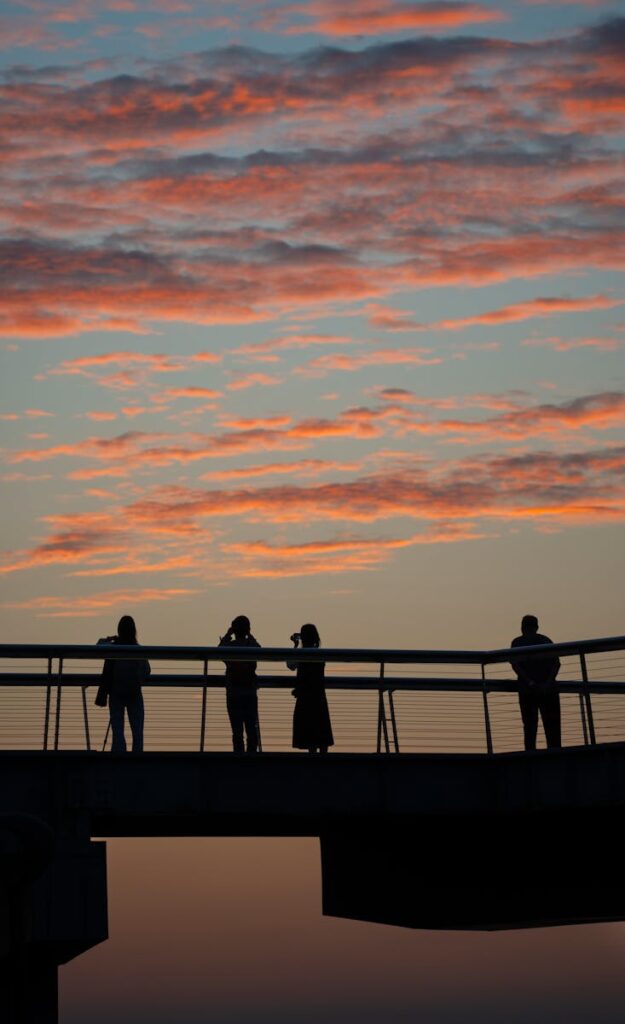 Silhouetted people standing on a footbridge against a colorful sunset sky.