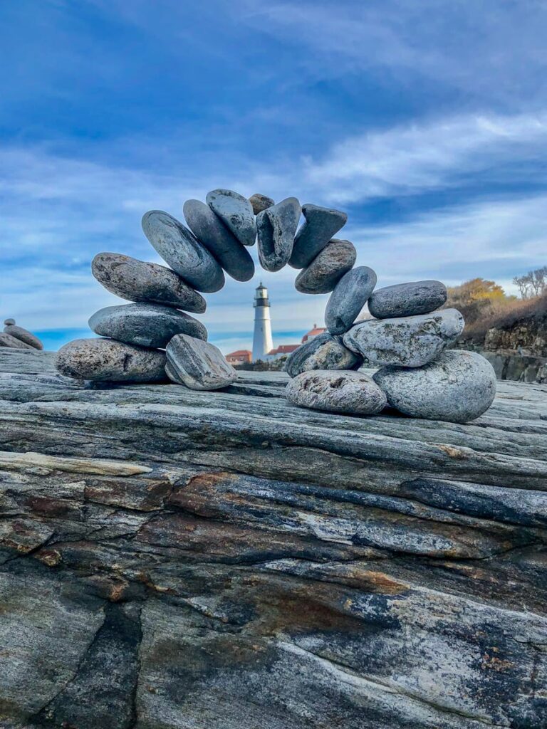 A serene image of balanced stones with the iconic Portland Head Lighthouse in the background.
