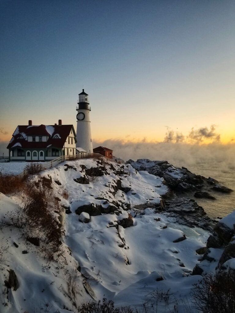 A serene winter morning at a snowy lighthouse on a rocky coast during sunrise.