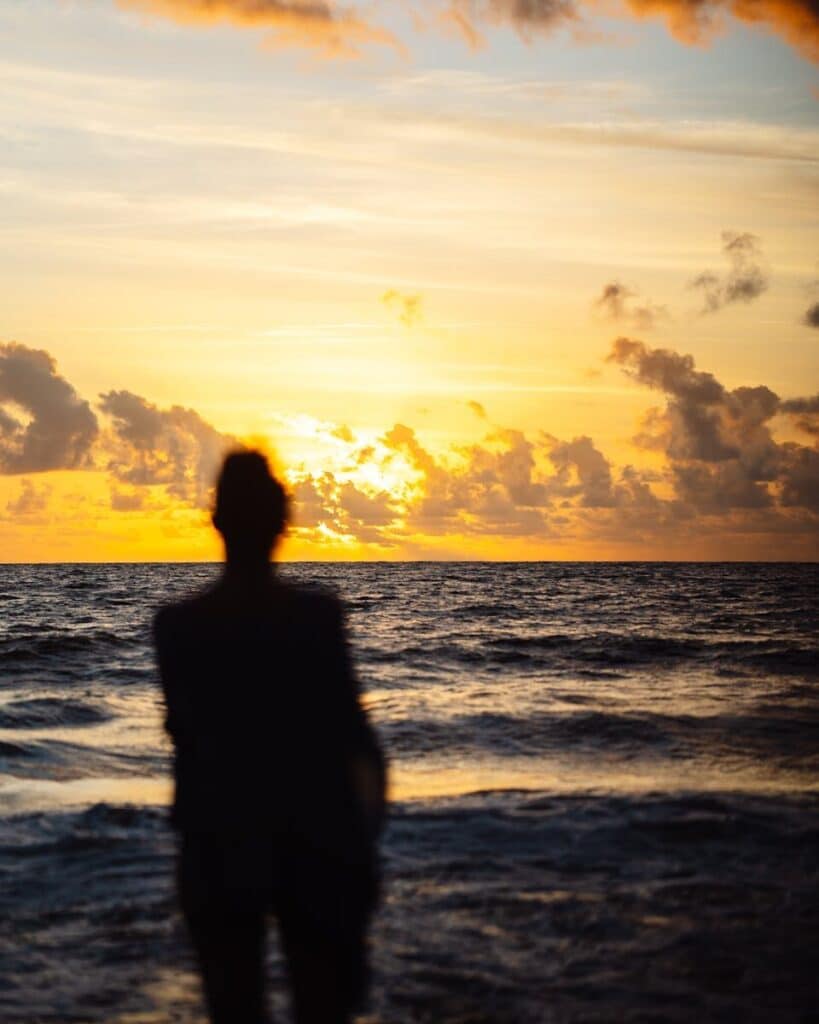 A silhouette stands by the ocean, admiring a beautiful orange sunset.